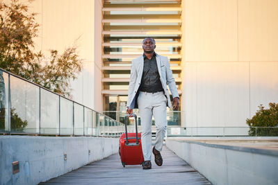 African-american man in a stylish suit and a red suitcase walking down a wooden walkway at sunset