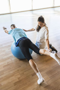 Fitness instructor helping athlete to balance on sports ball in gym