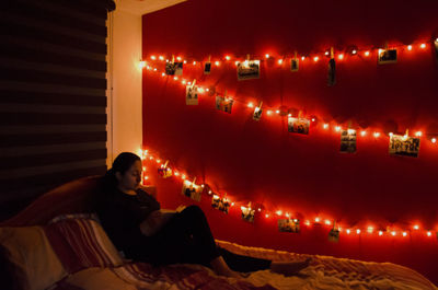 Full length of young woman sitting one bed with illuminated string lights and photographs against red wall in bedroom