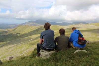 Rear view of friends sitting on grass while looking at landscape against cloudy sky