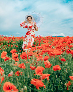Happy woman in summer dress with a umbrella dances in the field with blooming poppies against a sky