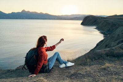 Woman sitting on rock at beach against sky during sunset