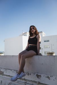 Low angle portrait of woman sitting on wall against clear sky