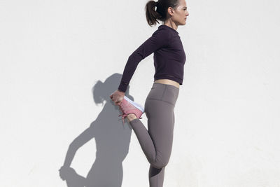 Fit sporty woman doing leg stretching exercises outside against a white wall.