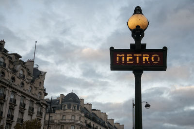 Low angle view of illuminated paris metro sign against cloudy sky