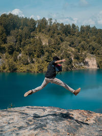 Woman jumping on rock by lake against trees