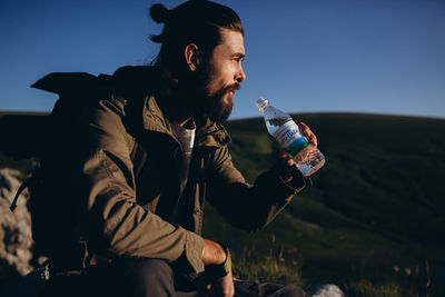 Young man drinking water from bottle while sitting against sky