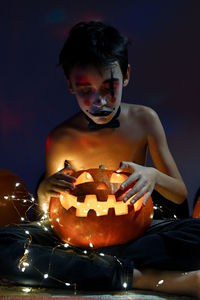 Full length of boy sitting by illuminated pumpkin during halloween