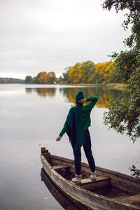 Woman in a green sweater stands with her back in an old wooden boat on a lake in autumn