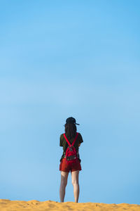 Rear view of backpack woman standing on sand against clear blue sky