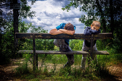 Bored friends leaning on wooden bench by trees against cloudy sky