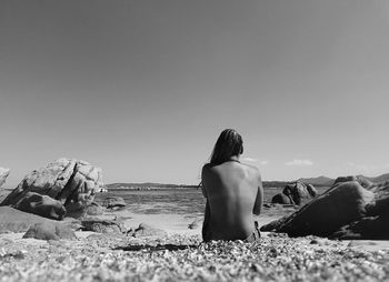 Rear view of topless sensuous woman sitting at beach against sky