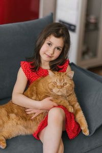Child girl holds her pet red british cat in her arms and sits with him on the couch at home