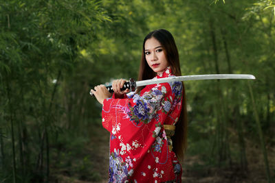 Portrait of young woman holding sword while standing in forest