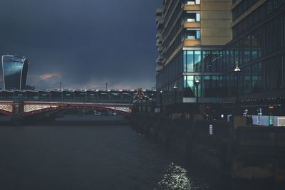 Illuminated bridge over river by buildings against sky at night