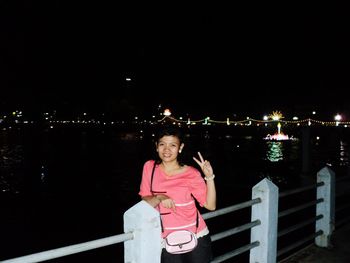 Portrait of woman standing by railing against river at night