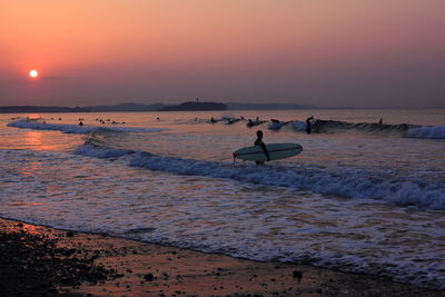 Surfer walking at beach against orange sky