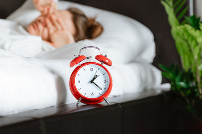 Young woman waking up in white bed yawning rubbing eyes turning alarm clock in bedroom. 