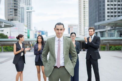 Portrait of young businessman standing against colleagues clapping on footpath against buildings in city