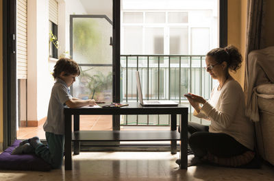 Side view of woman working while son studying at table during lockdown