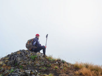 Man sit with a backpack and north walking poles on top of a rock against mist in the mountains. 