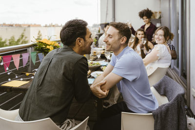 Romantic gay couple looking at each other while sitting with friends at dinner party in balcony