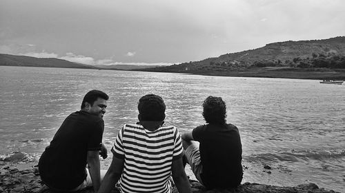 Rear view of friends sitting on shore at beach against sky