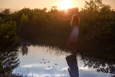 Rear view of woman standing by lake against sky during sunset