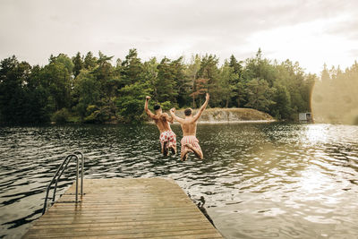 Male friends with arms raised jumping in lake from jetty during vacation