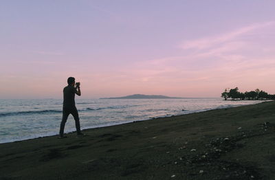 Silhouette man photographing at beach while walking against cloudy sky