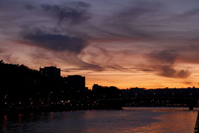 Silhouette buildings by river against sky during sunset