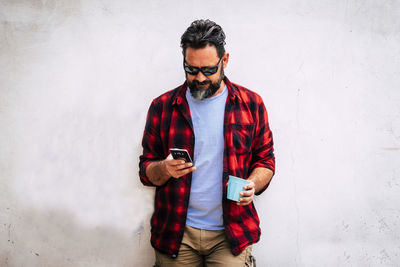 Young man using phone while standing against wall