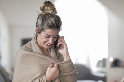 Woman wrapped in blanket talking on mobile phone while standing at home