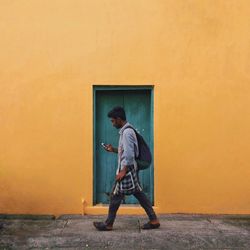 Side view of man using mobile phone while walking against yellow wall