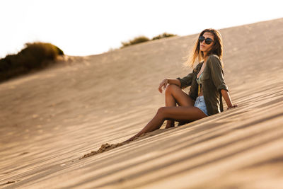 Young woman sitting on sand against sky