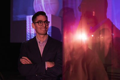 Smiling male entrepreneur with arms crossed standing at illuminated exhibition center