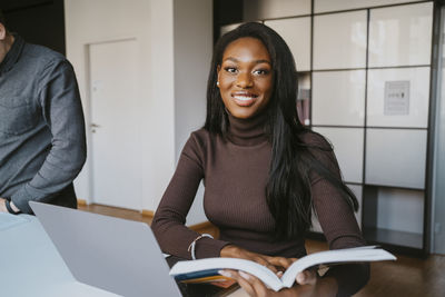 Portrait of smiling young female student sitting with book studying by friend at community college