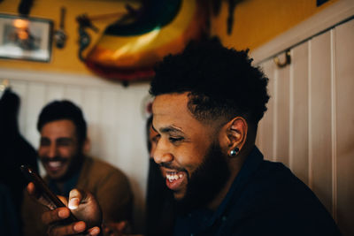 Smiling young man using smart phone while sitting by multi-ethnic friends in restaurant