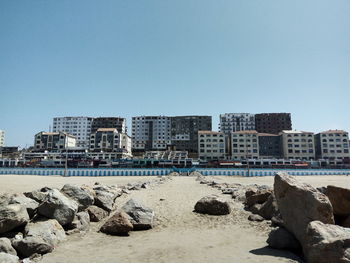 Rocks at sandy beach by buildings against clear sky