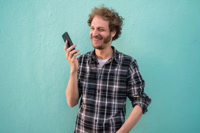 Smiling young man using mobile phone against wall