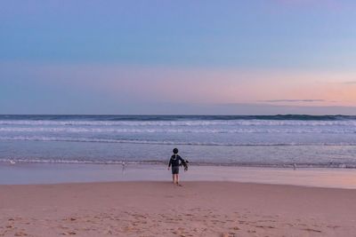 Rear view of boy carrying seaweed on sea shore against sky during sunset