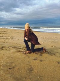 Full length of woman kneeling on sea shore at beach against cloudy sky