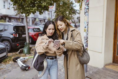 Young female friends sharing smart phone while standing on sidewalk