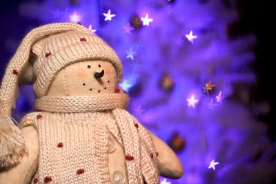 Close-up of woolen snowman with illuminated christmas tree in background