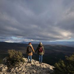 Rear view of friends standing on mountain against cloudy sky during sunset