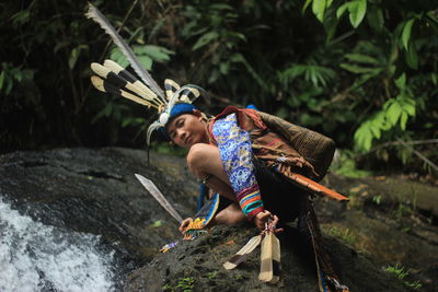 Low angle portrait of young man wearing traditional clothing crouching on rock against plants