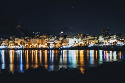 Illuminated buildings by sea against sky at night