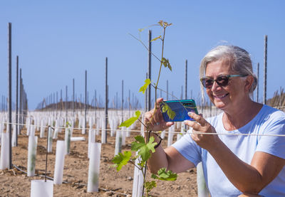 Smiling woman photographing plant while standing against sky