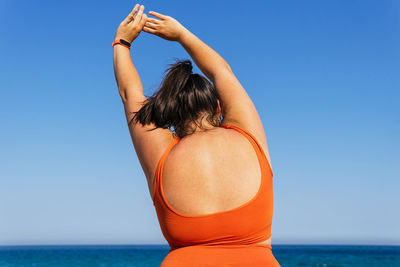 Back view of anonymous plump female athlete in sportswear exercising with raised arms against ocean under blue sky