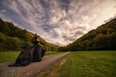 Rear view of woman wearing black dress on road against cloudy sky during sunset